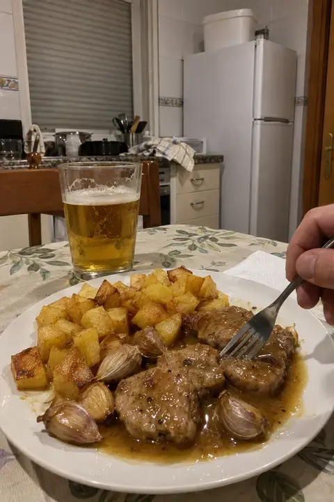 Plato de solomillo al whisky con ajos enteros y patatas en la mesa de una cocina, con un tenedor en la mano